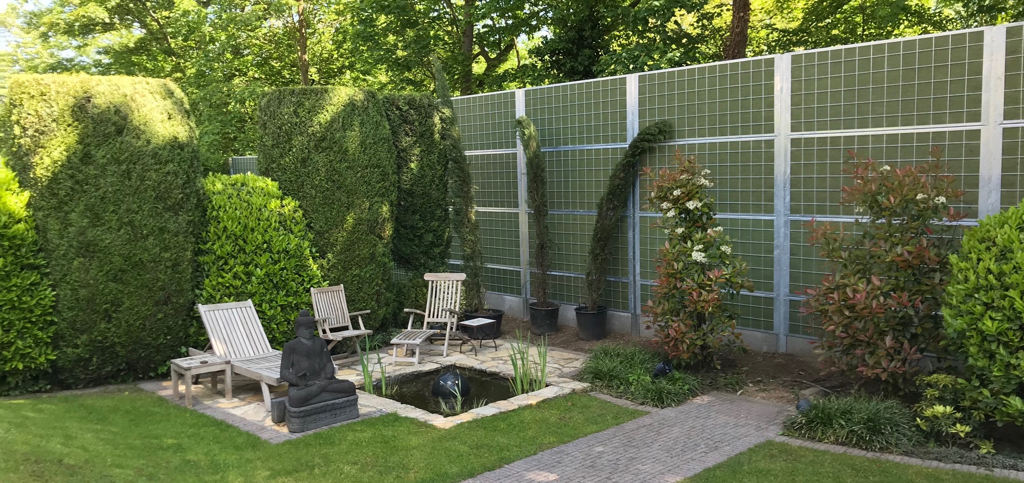 En mysig trädgård med buskar och fiskdamm, har ett Noistop® Steel bullerplank från Poda Stängsel uppsatt som avskärmning till grannarna.