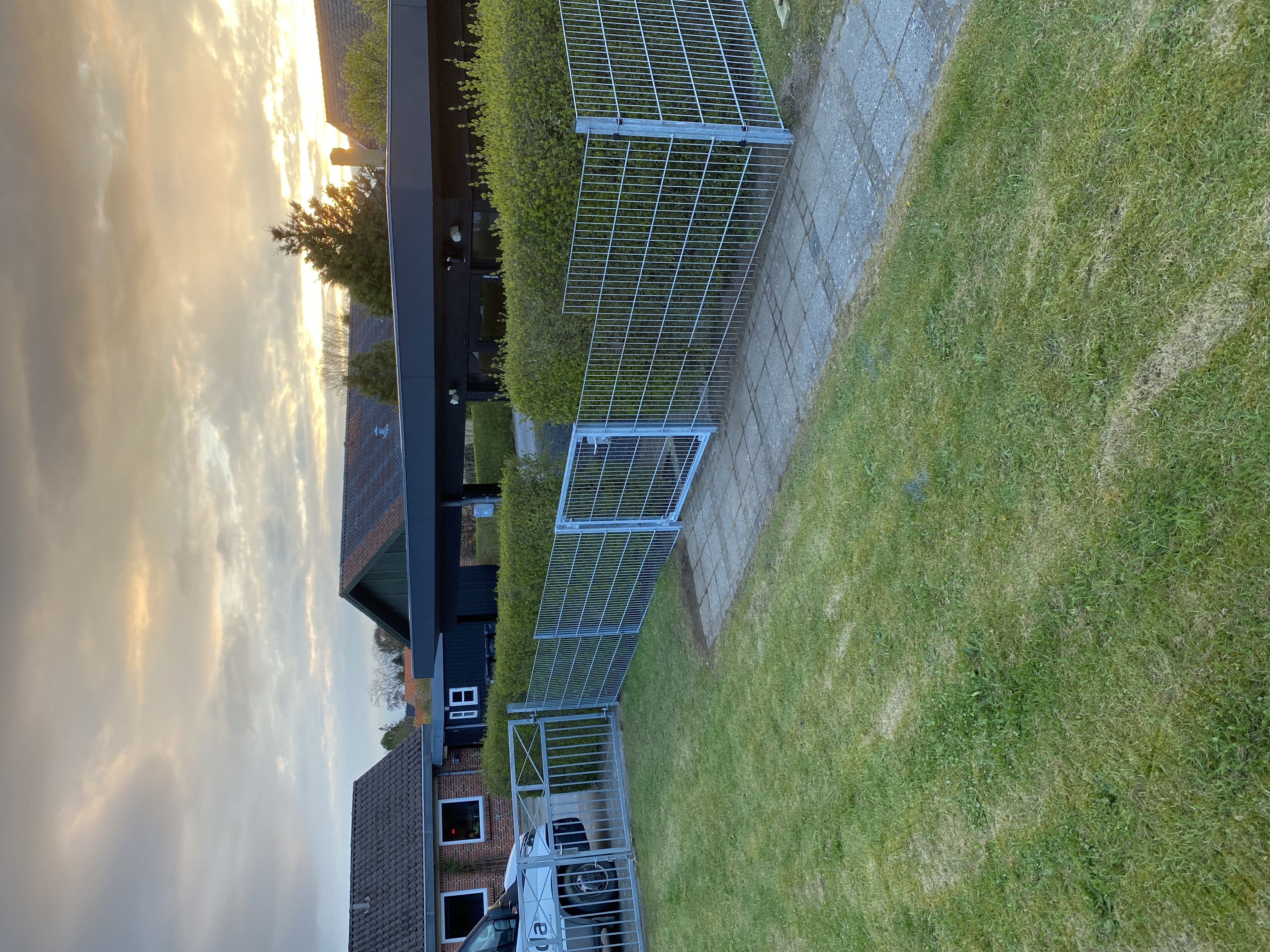 Panelstängsel med en grind monterad längs en stig som skiljer en gräsmatta från en häck. I bakgrunden syns en carport, flera hus och en solnedgång.